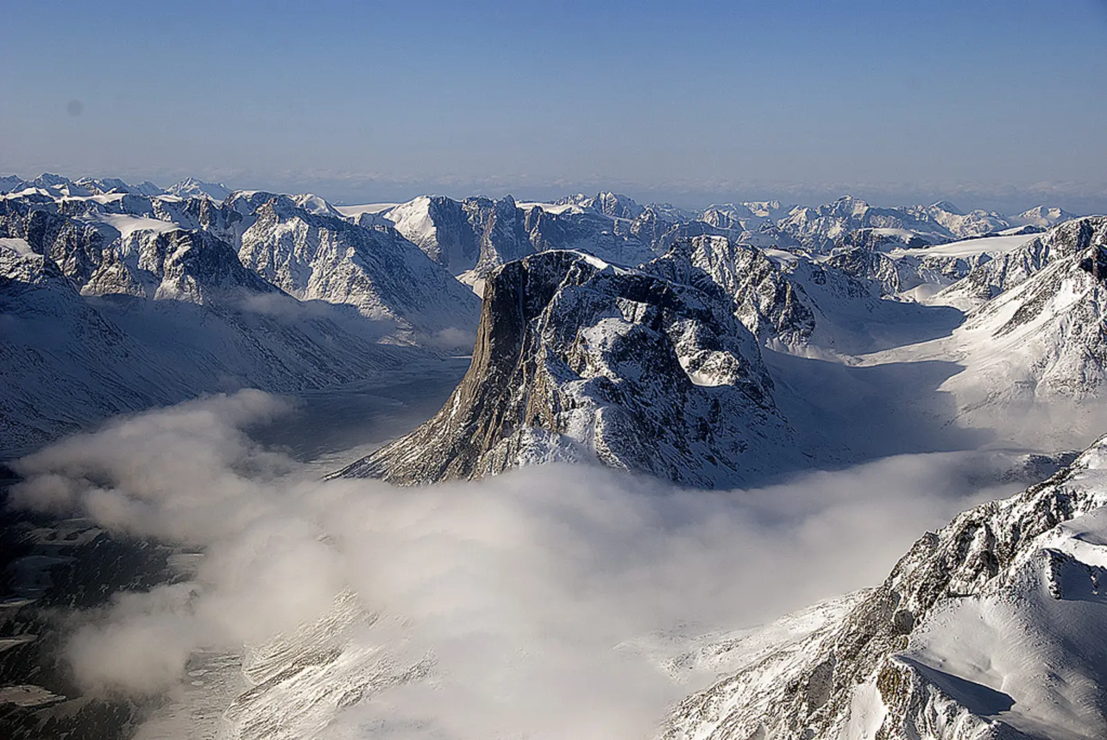 A snow-covered mountain peak rises above a thick layer of low-lying clouds, surrounded by a vast, jagged mountain range under a clear blue sky. The photographer titled it “SW Greenland Fjord and Mountain”.