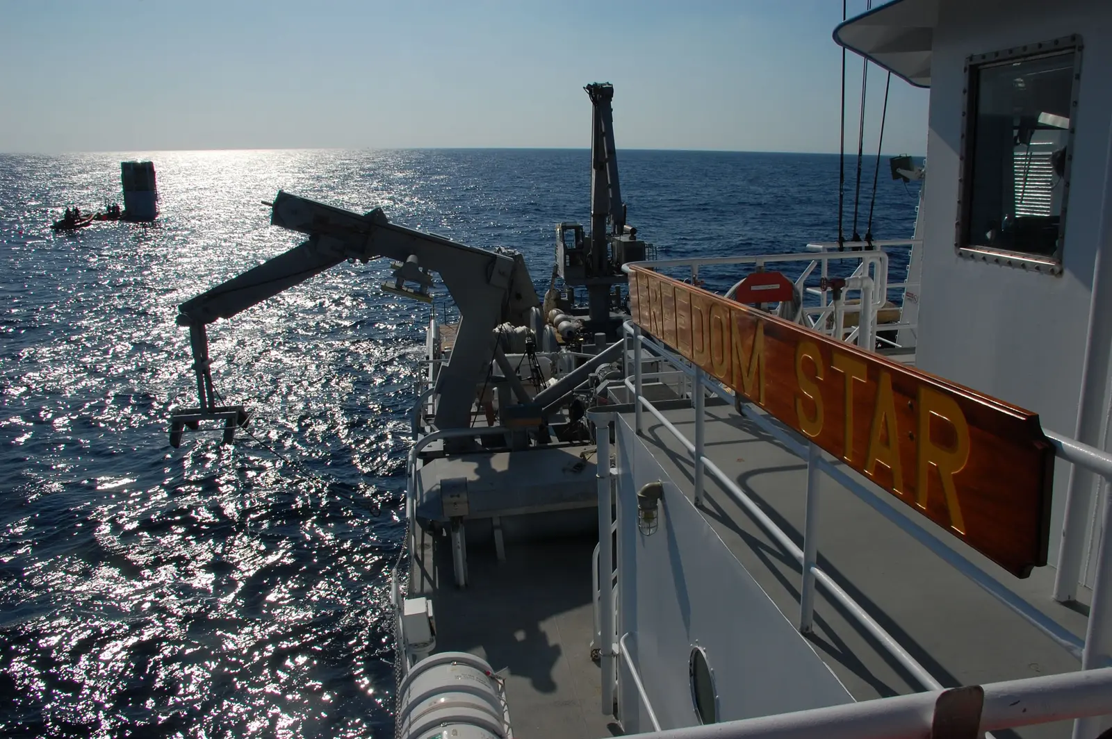 A view from the deck of a ship at sea, showing a mechanical crane arm extending over the water and a wooden sign reading "DOM STAR".