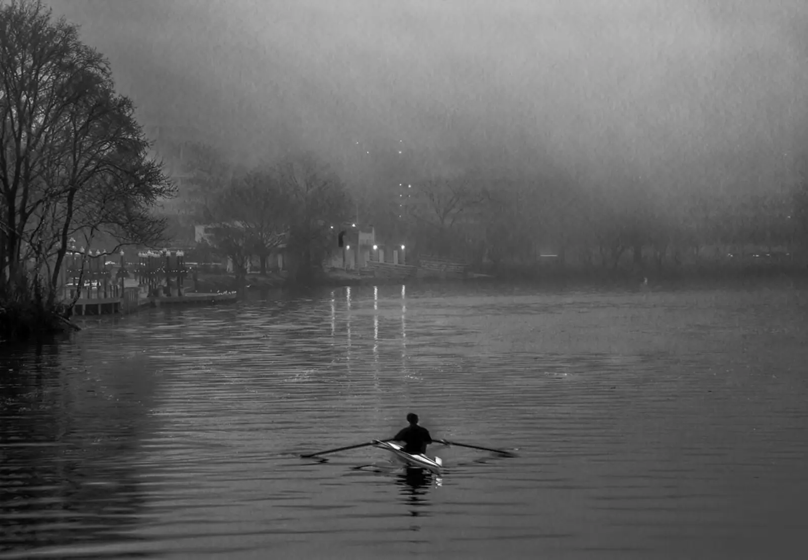 A solitary figure rows a boat across a wide, mist-covered body of water in a black-and-white image. The photographer titled it “Foggy Bottom”.