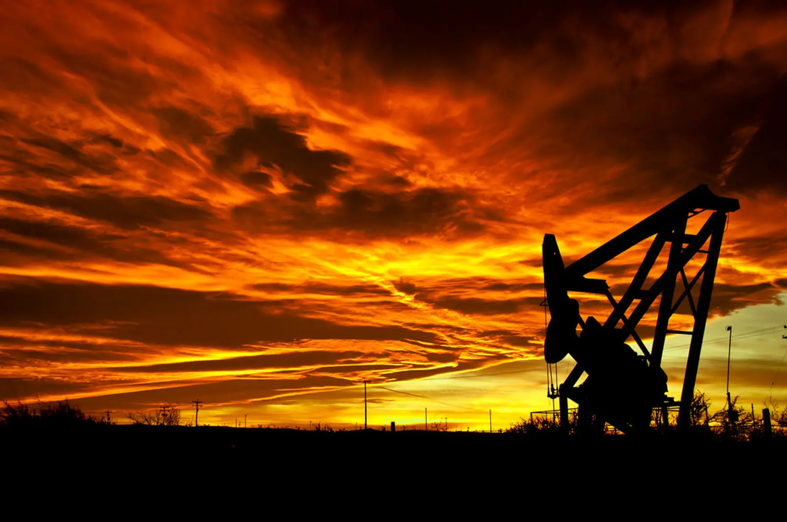 An oil pump jack is silhouetted against a dramatic, fiery orange and yellow sunset sky. The photographer titled it “The Burning of the Sky”.