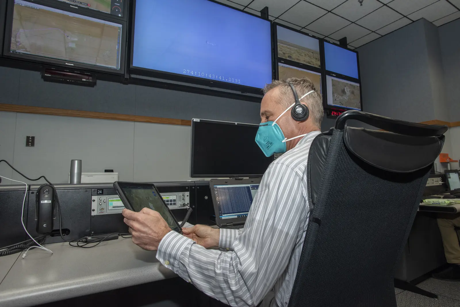 A man wearing a headset and a teal face mask sits in a control room, looking at a tablet while multiple large screens display maps and data in the background. The photographer titled it “TG-14 Flight Tests Support the Advanced Air Mobility Project's National Campaign”.