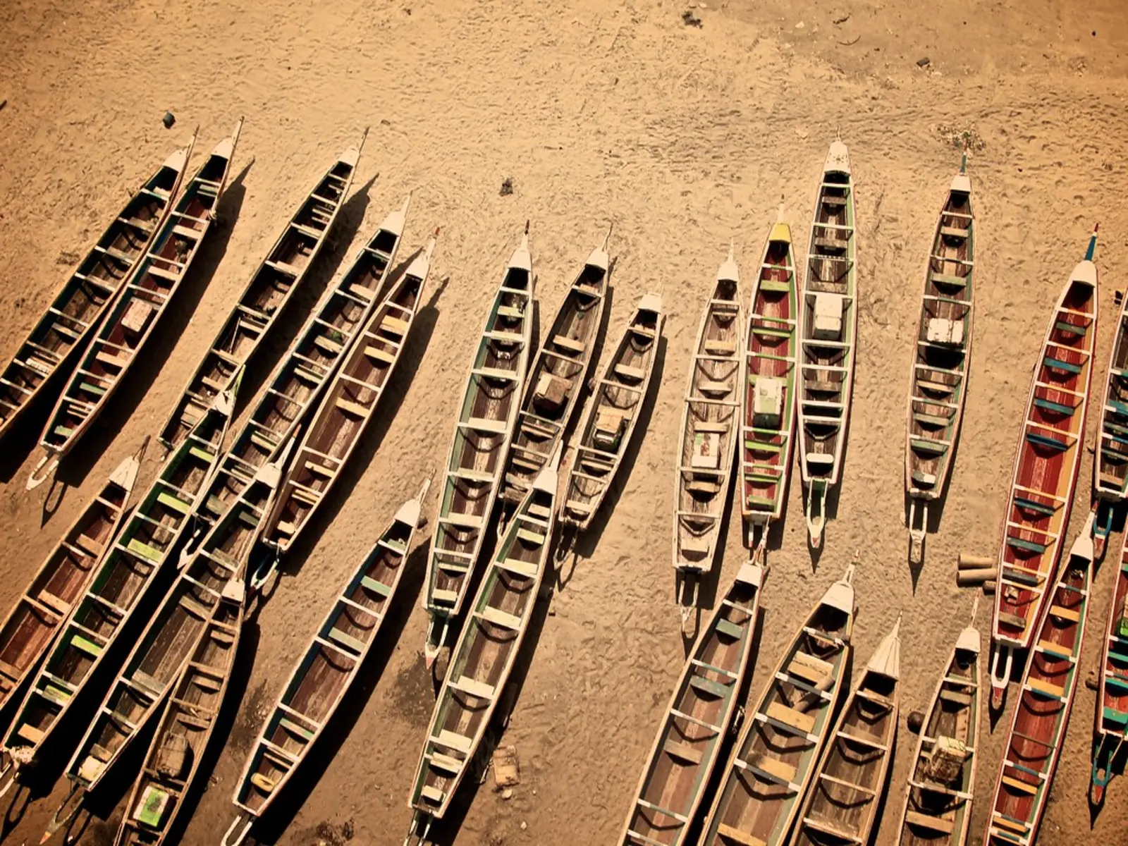 An aerial view shows numerous empty, narrow wooden boats resting on a sandy beach. The photographer titled it “Soumbedioune Boats”.