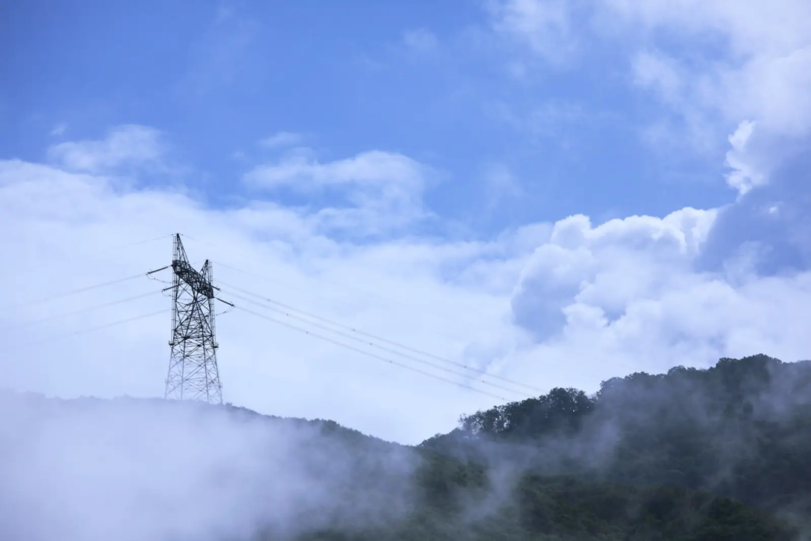 A steel lattice transmission tower stands on a misty, tree-covered ridge against a blue sky filled with white clouds. The photographer titled it “Transmission Tower / 鉄塔(てっとう)”.