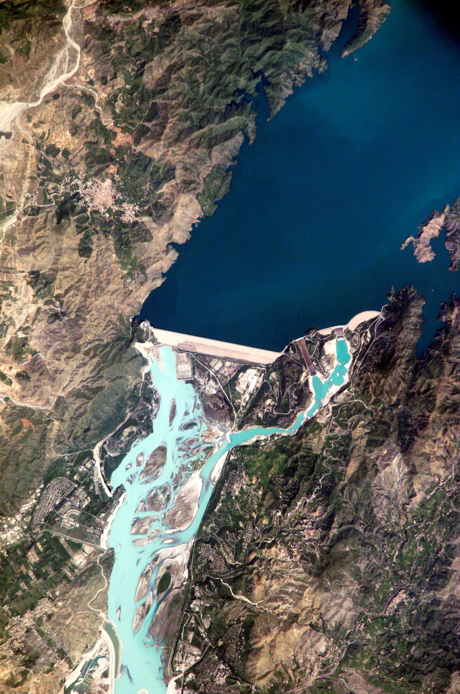 A satellite photograph shows a large dam holding back a deep blue reservoir, with a turquoise river flowing through a mountainous landscape below. The photographer titled it “Crew Earth Observations (CEO) taken during Expedition Five on the ISS”.