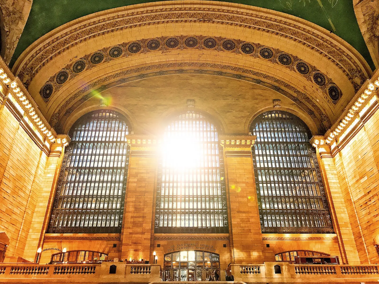 The interior of a grand terminal hall shows three tall arched windows set beneath an ornate vaulted ceiling decorated with rows of circular medallions, with bright sunlight flaring through the central window. The photographer titled it “Grand Central Terminal Train Station in New York City”.