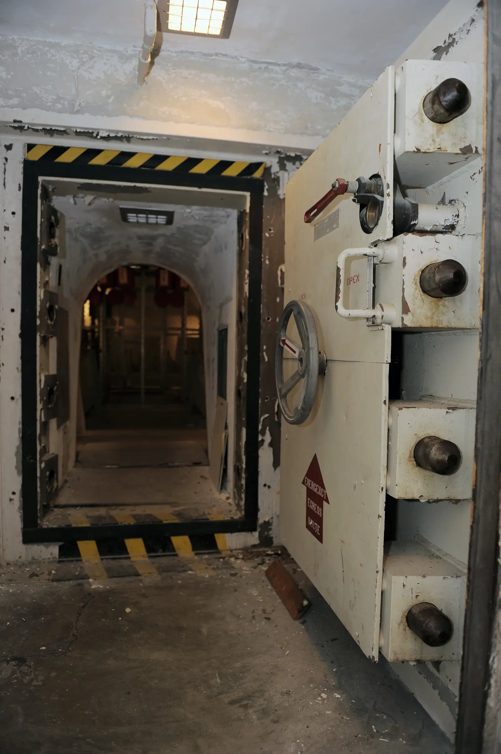 A large, white metal bank vault door with heavy locking bolts stands open, revealing a dark, arched hallway beyond.