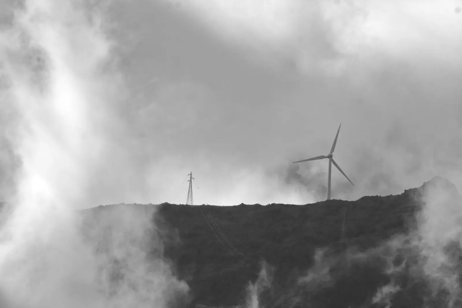 A black and white photograph shows a wind turbine and a transmission tower silhouetted against a cloudy sky on a dark, sloping ridge. The photographer titled it “Wind Turbine & Clouds”.
