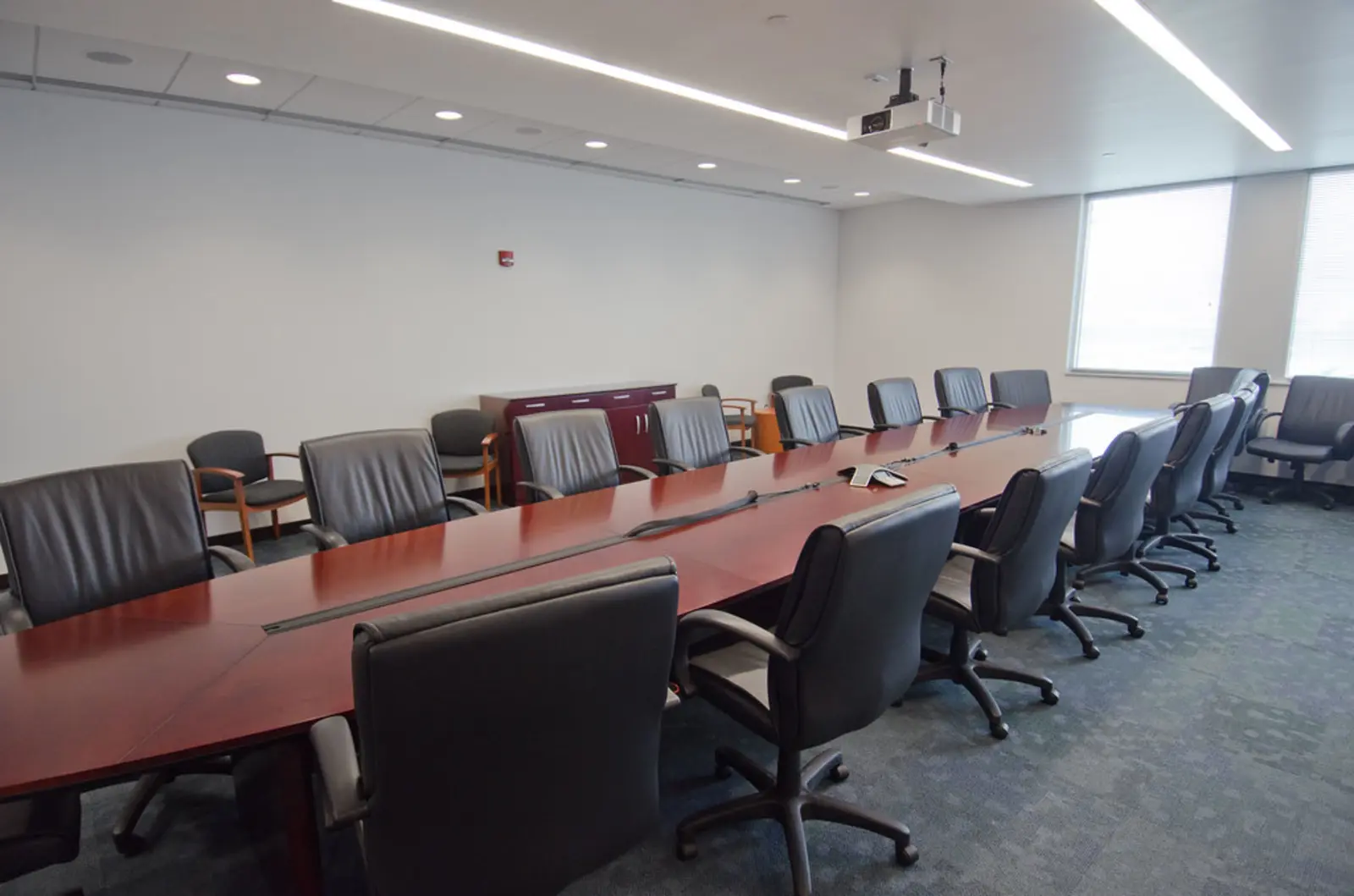 A long, reddish-brown wooden conference table is surrounded by empty black leather office chairs in a brightly lit room featuring a ceiling-mounted projector and large windows.