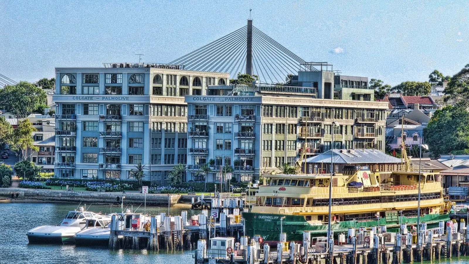 A large, light-colored factory building displaying the name 'Colgate-Palmolive' stands beside a river, where a yellow and green ferry is docked at a pier. The photographer titled it “Colgate-Palmolive Factory”.