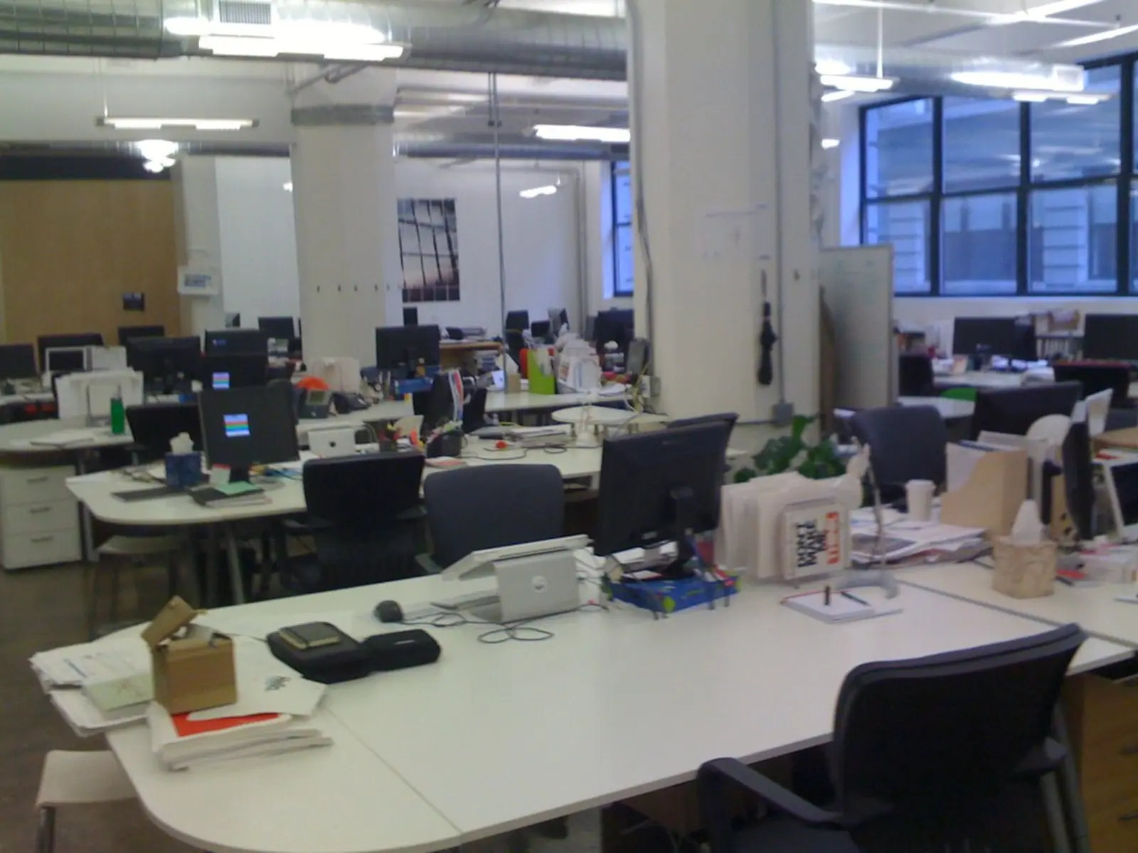 An open-plan office space is shown with rows of white desks, multiple computer monitors, and black office chairs.