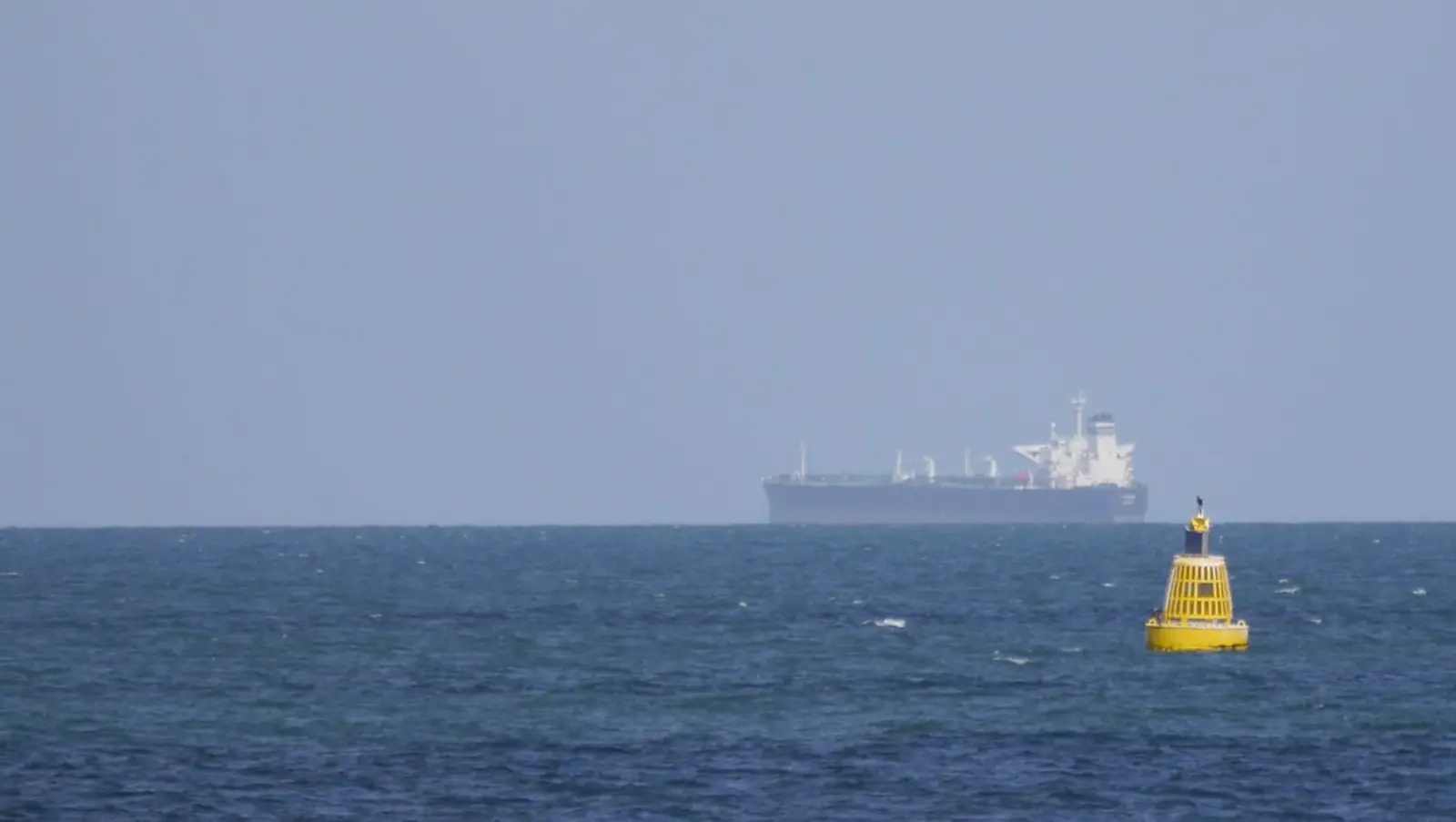 A large cargo ship is visible on the horizon of the open sea, with a yellow buoy floating in the foreground. The photographer titled it “Distant Cargo Vessel”.