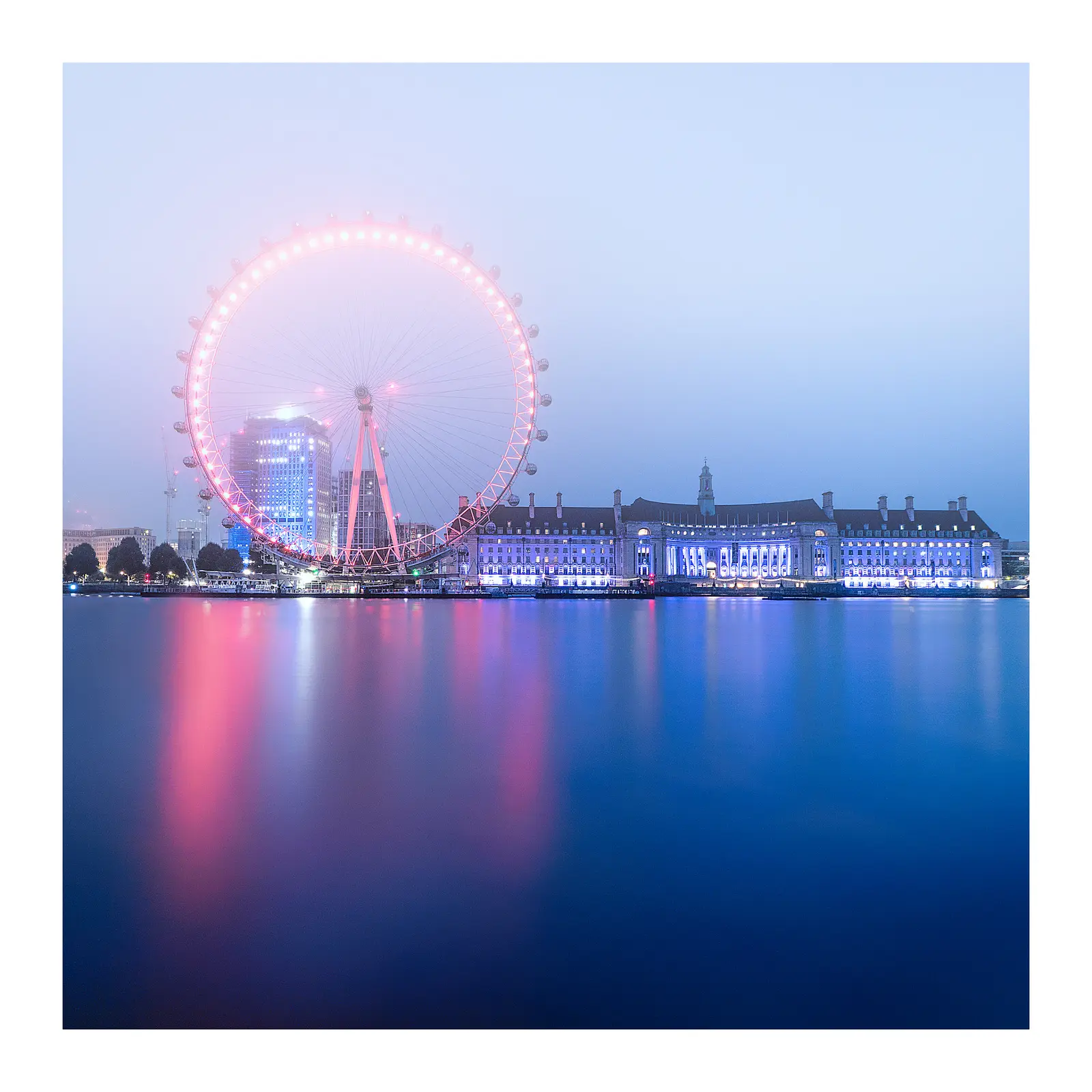 A large observation wheel glowing pink and a long building illuminated in blue reflect on the calm water of a river under a hazy sky. The photographer titled it “<div class='fn'> London Eye</div>”.