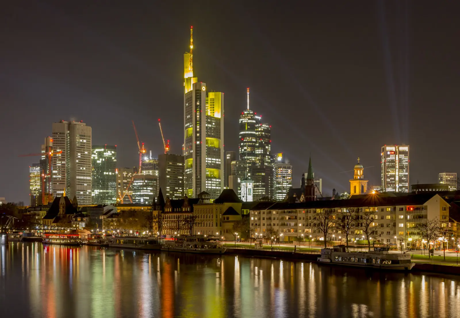 Illuminated skyscrapers and historic buildings line the edge of a river at night, their lights reflecting on the water's surface. The photographer titled it “Frankfurt at night”.