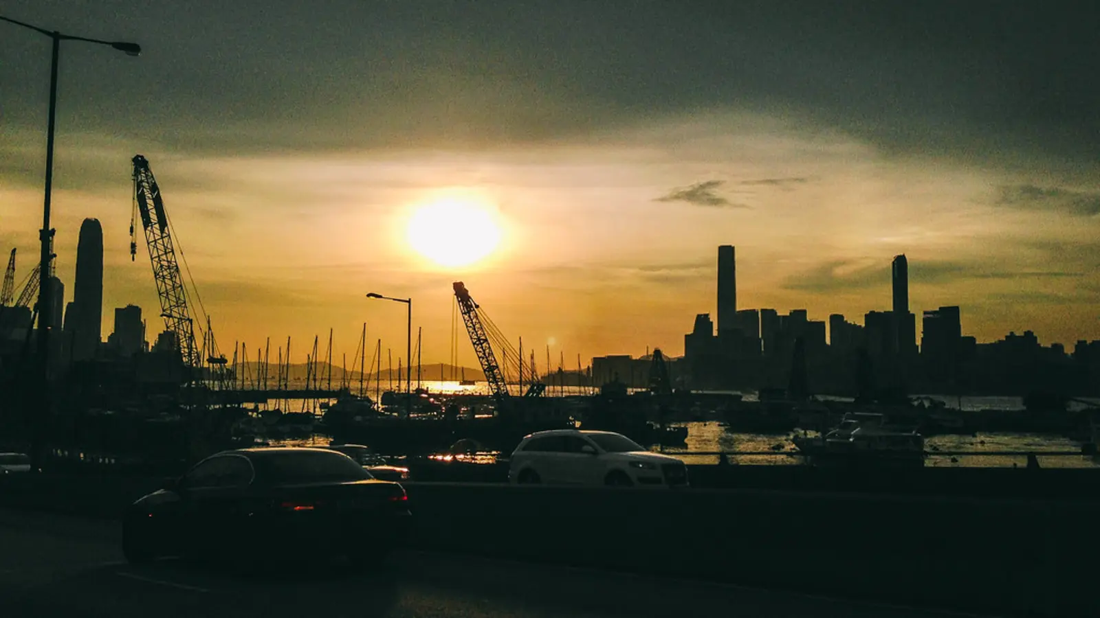 A sunset over a city skyline featuring construction cranes, boats, and cars on a road in the foreground. The photographer titled it “Hong Kong Victoria Harbour Sunset”.