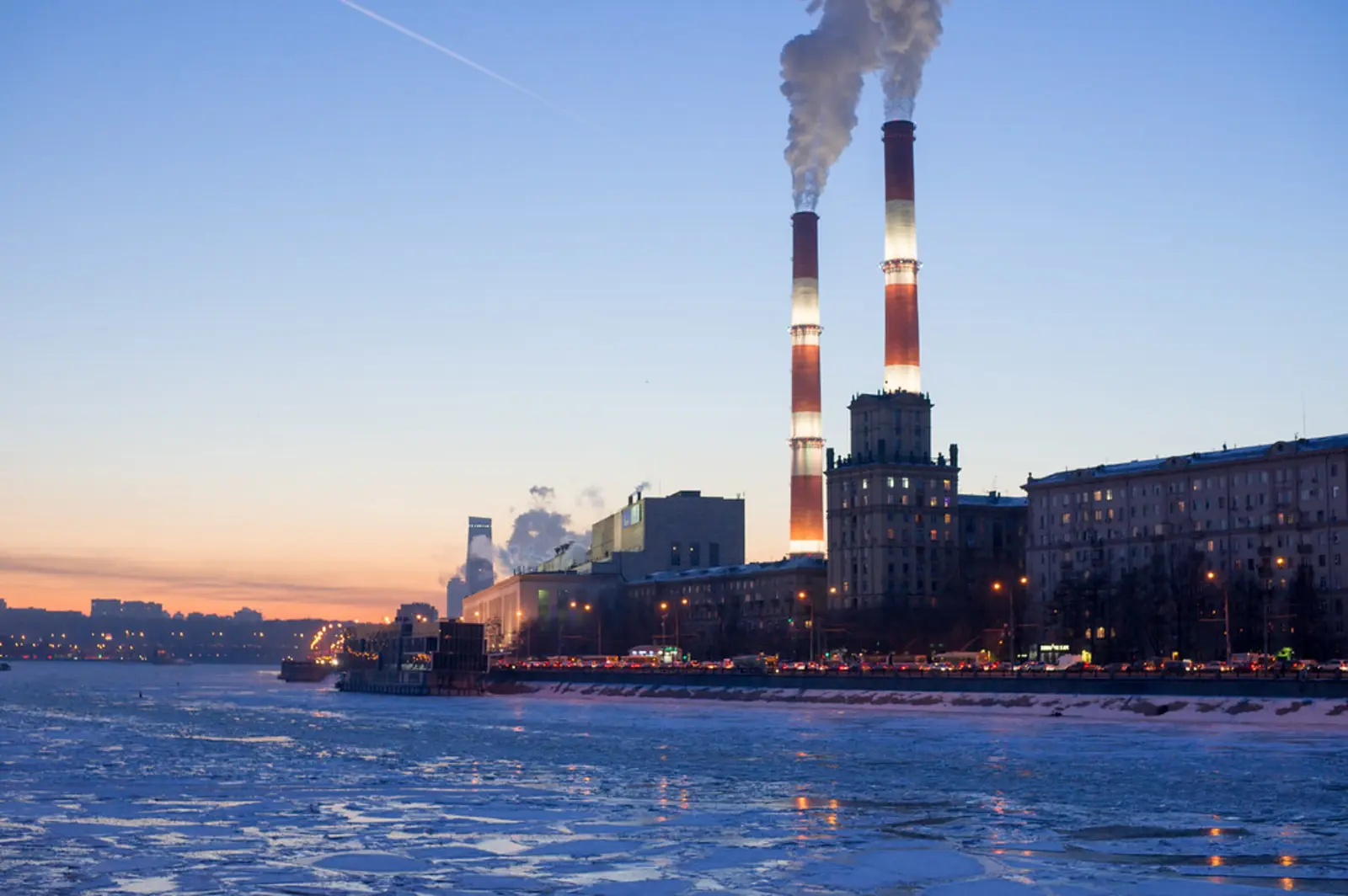 A large power plant with two tall, red-and-white striped smokestacks emitting smoke stands beside a frozen river under a twilight sky. The photographer titled it “Power plant №12 in winter”.