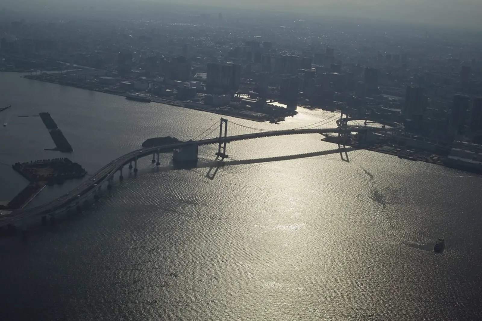 An aerial view shows a long bridge crossing a body of water towards a hazy city skyline, with bright sunlight reflecting off the surface. The photographer titled it “Tokyo Bay Bridge from Above”.