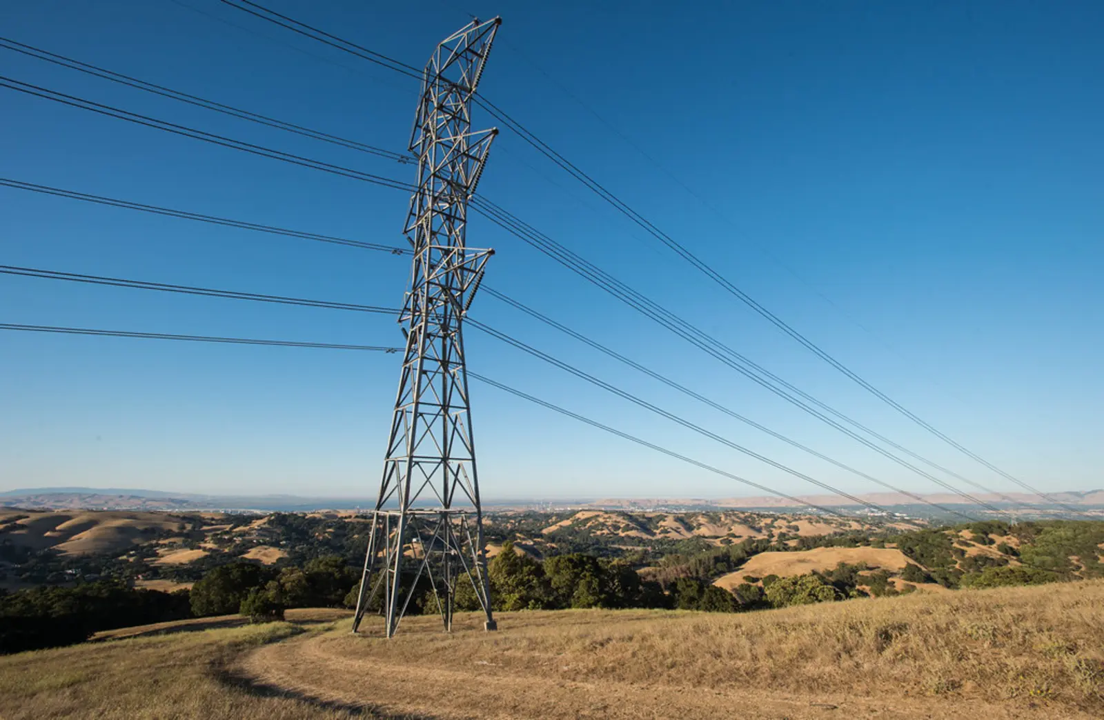 A tall metal electrical transmission tower stands on a grassy hill, carrying power lines across a hilly landscape under a clear blue sky.
