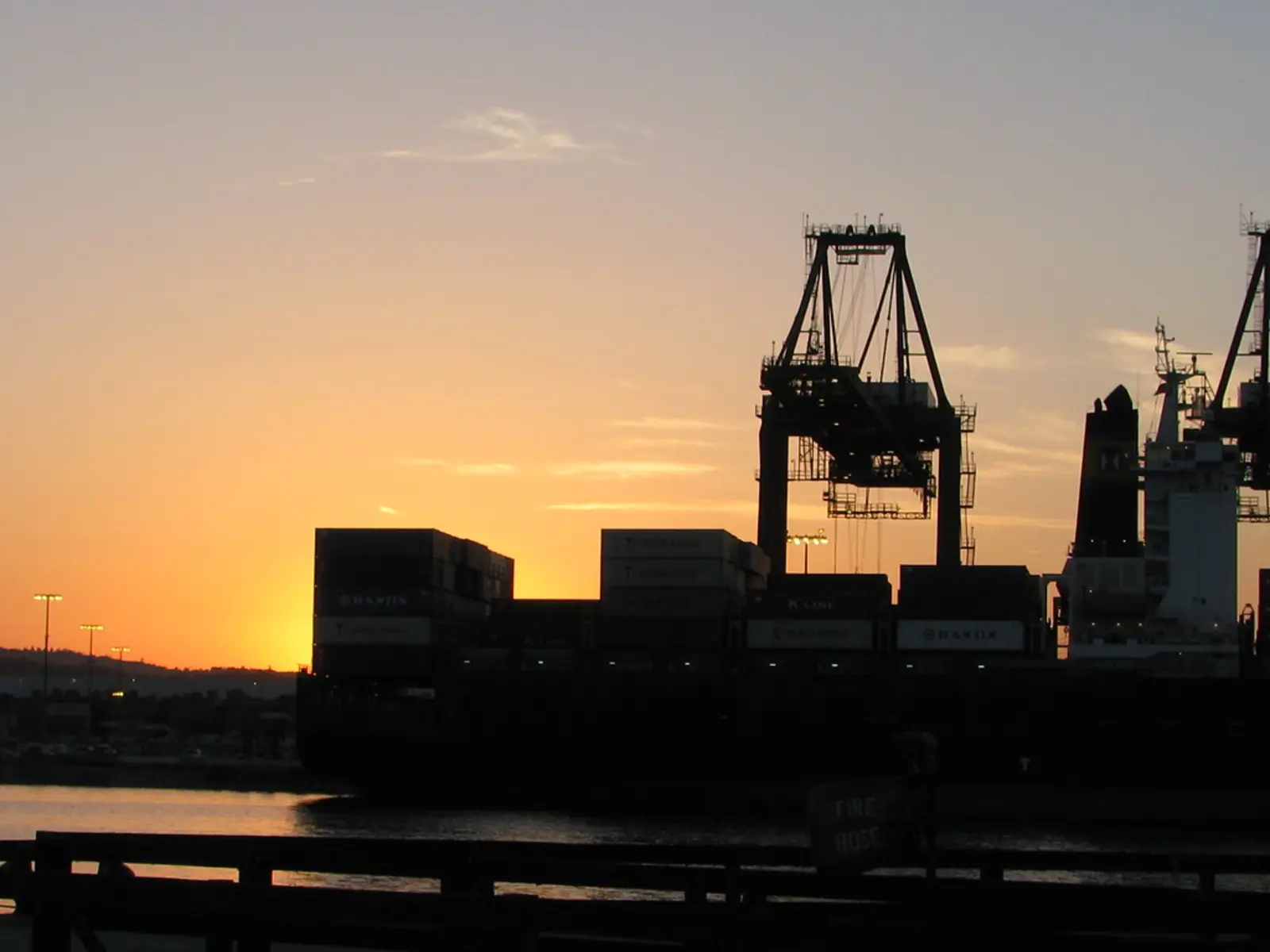 A large container ship sits silhouetted at a port terminal during sunset, with a loading crane visible nearby.