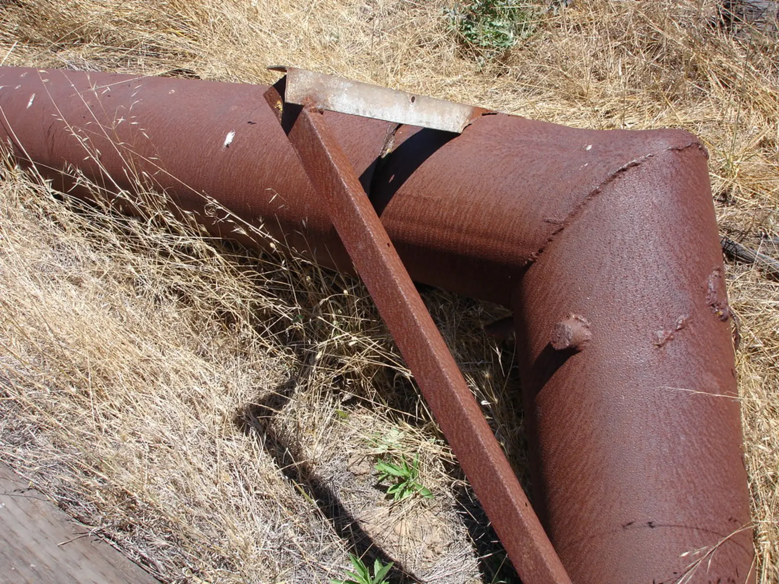 A large, heavily rusted section of an oil pipeline lies in dry grass next to a bent, weathered metal support bracket.